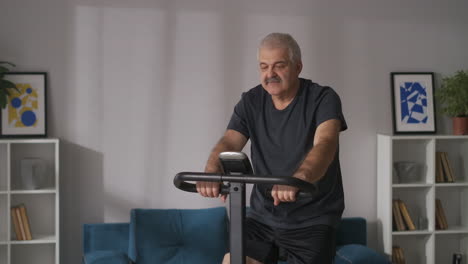 Retrato-De-Un-Hombre-De-Mediana-Edad-En-Bicicleta-Estática-En-Un-Apartamento-Entrenando-En-Casa-Con-Ejercicios-Cardiovasculares-Para-Mantener-Una-Buena-Condición-Física